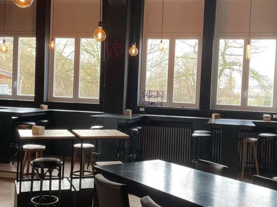 De zaal met hoge ramen, staantafels en hanglampen.